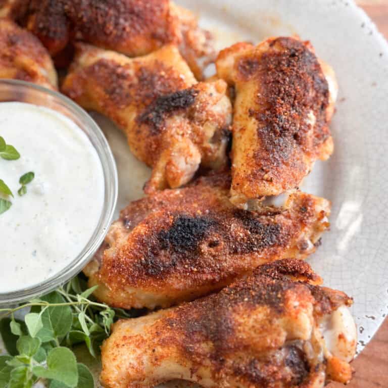 A plate of seasoned and grilled chicken wings is shown next to a small bowl of creamy dipping sauce and a sprig of fresh herbs on a white, textured plate.