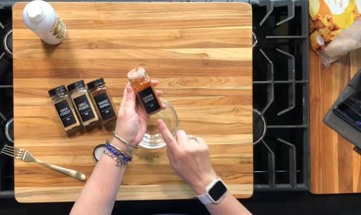 A person holding a small jar of ground cayenne pepper above a wooden cutting board, which has jars of garlic powder, onion powder, and ground pepper, a fork, and other kitchen items on it.