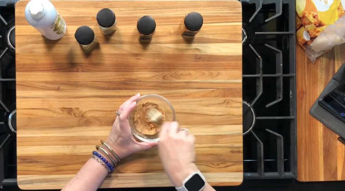A person mixes brown seasoning in a glass bowl on a wooden cutting board. Four spice jars, a white spray bottle, and part of a bag of chips are visible nearby on a stove.