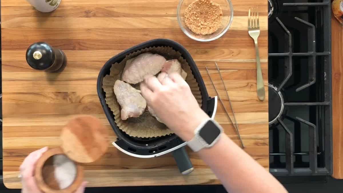 A person seasons raw chicken wings in an air fryer basket lined with parchment paper, with a pepper grinder, bowl of seasoning, utensils, and a stove visible on a wooden countertop.
