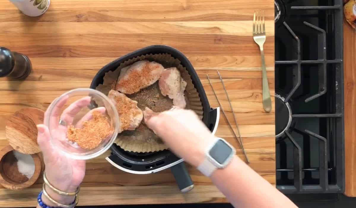 A person sprinkles seasoning from a small bowl onto raw chicken pieces arranged in an air fryer basket on a wooden countertop, with kitchen utensils and a stove nearby.
