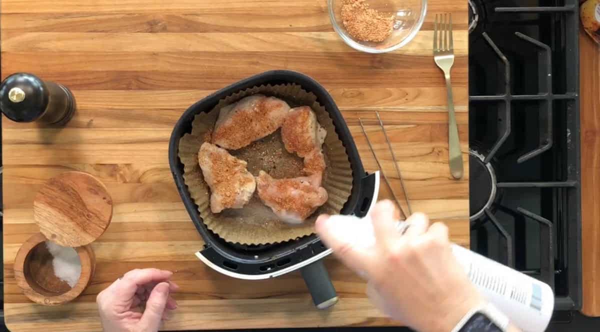 A person sprays oil over seasoned chicken pieces arranged on parchment paper in an air fryer basket. A bowl of seasoning, a salt cellar, a pepper grinder, and a stove are visible on the wooden countertop.