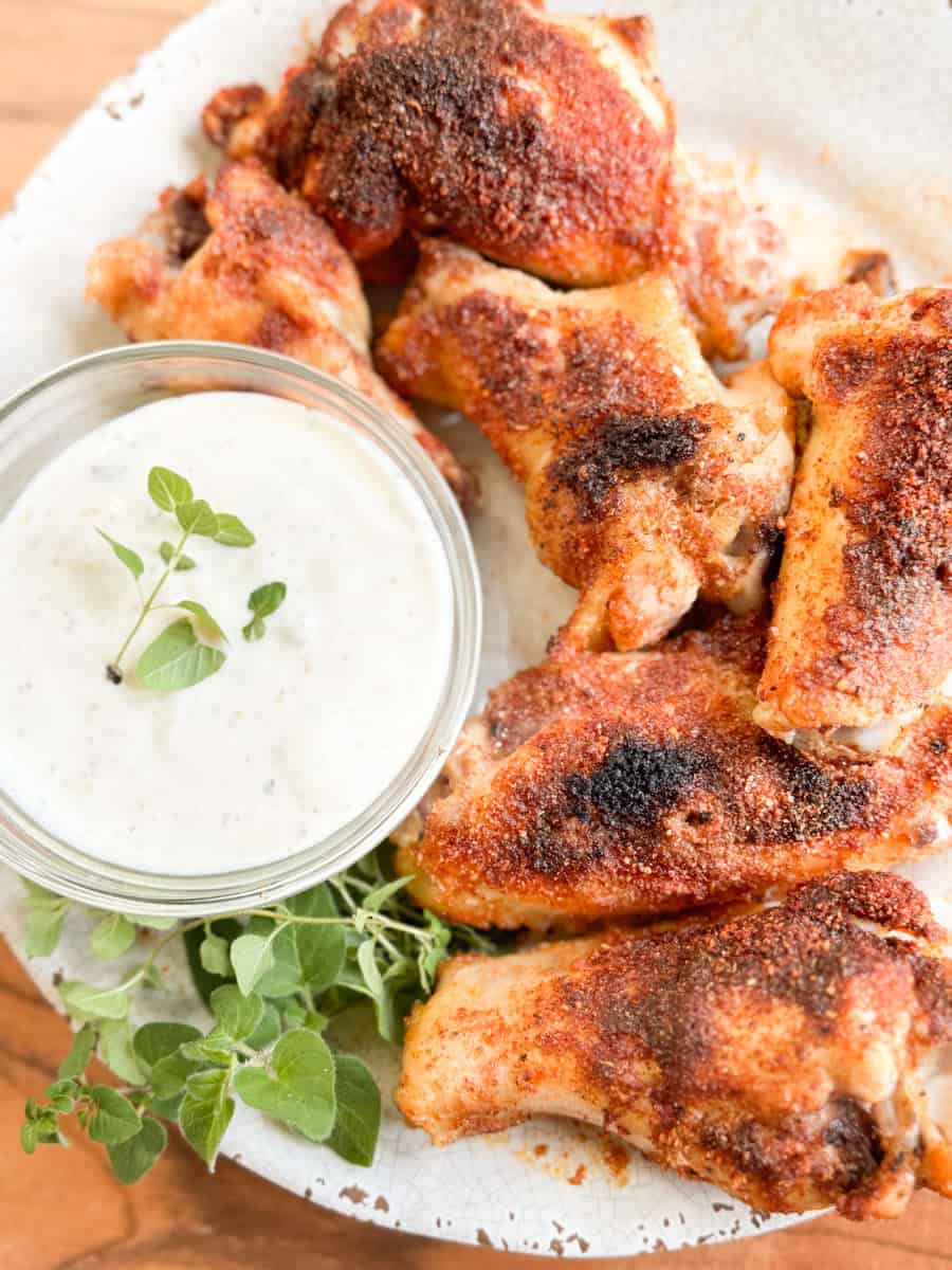 A plate of seasoned, baked chicken wings is arranged next to a small bowl of creamy dipping sauce, garnished with fresh herbs. The wings have a browned, crispy coating.