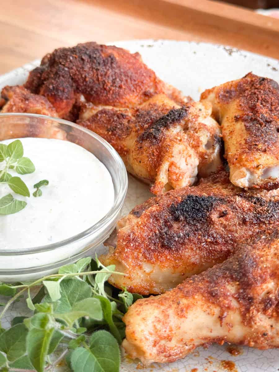 Four pieces of seasoned, cooked chicken are arranged on a plate next to a small glass bowl of creamy dipping sauce and a sprig of fresh oregano. The chicken has a crispy, browned exterior.