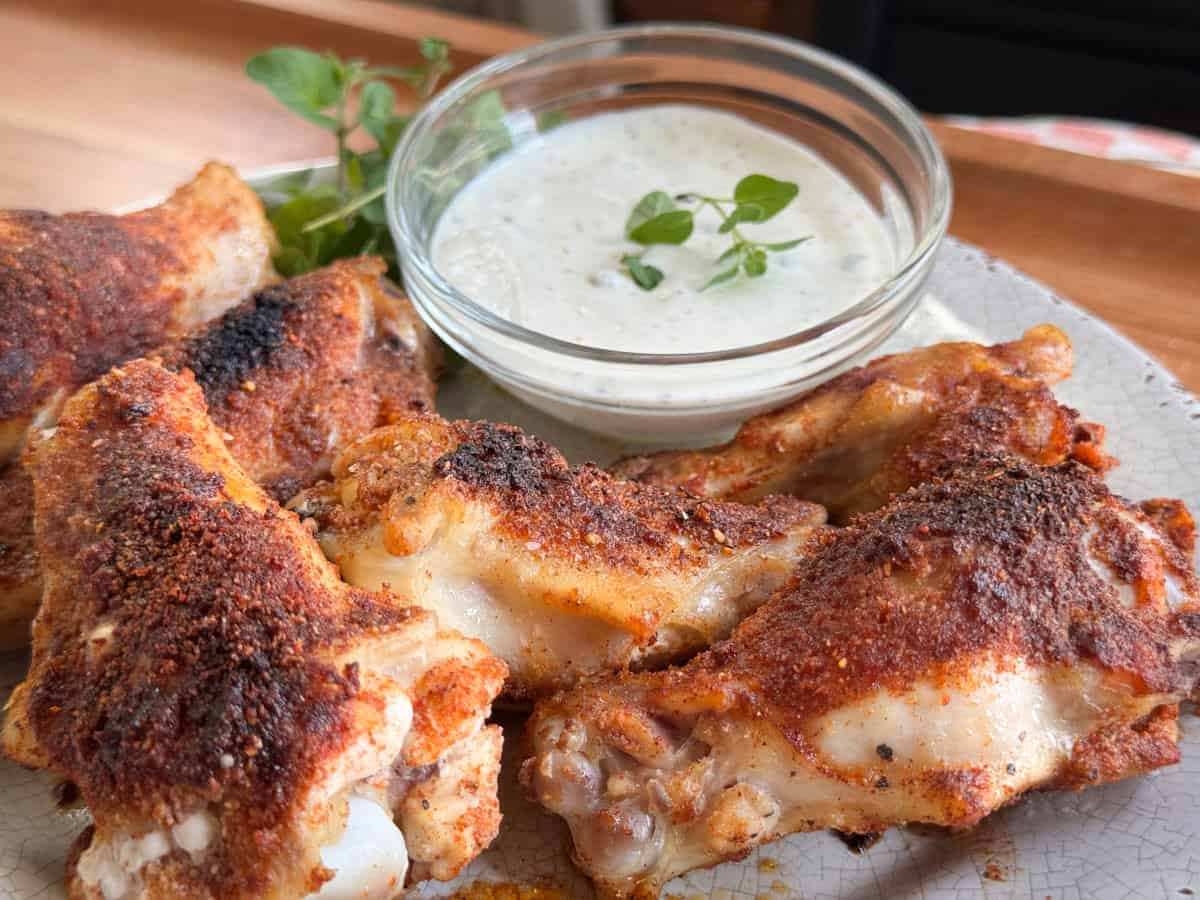 A plate of seasoned, baked chicken wings is arranged around a clear bowl of white dipping sauce, garnished with fresh herbs. The wings are browned and appear crispy, with visible spices on the surface.