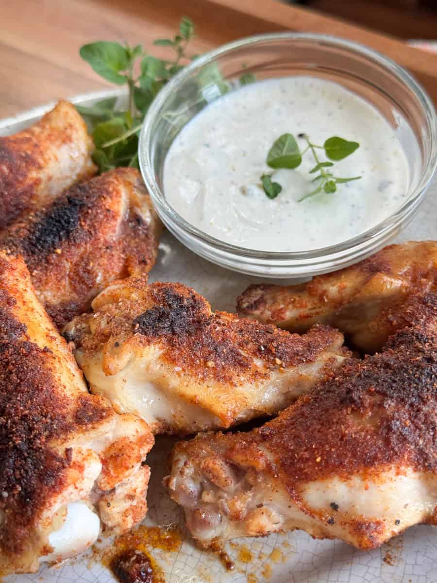 Four seasoned and baked chicken wings are arranged on a plate next to a small glass bowl of creamy dipping sauce, garnished with fresh herbs.