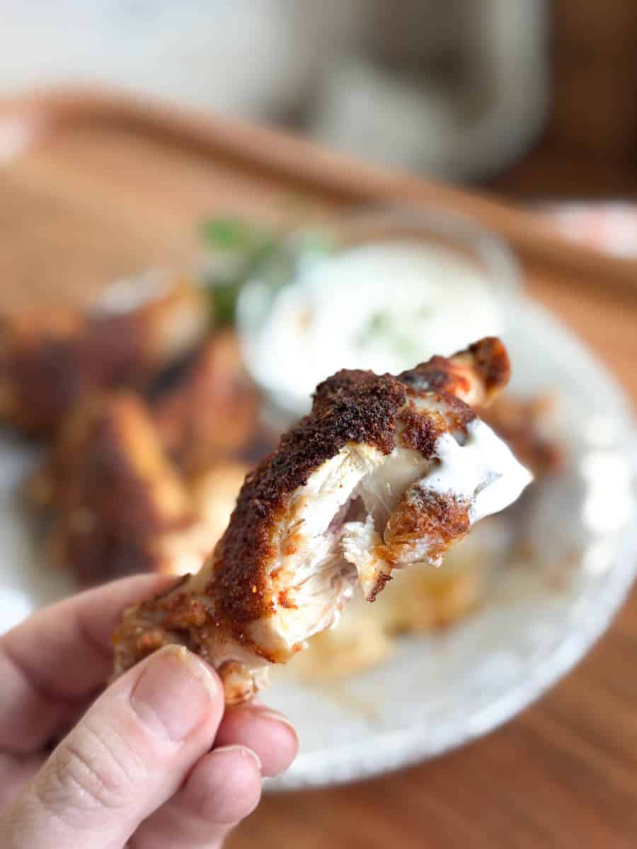 A hand holds a partially eaten, seasoned chicken wing with a bit of white dipping sauce on it. More chicken wings and a small bowl of sauce are on a plate in the blurred background.