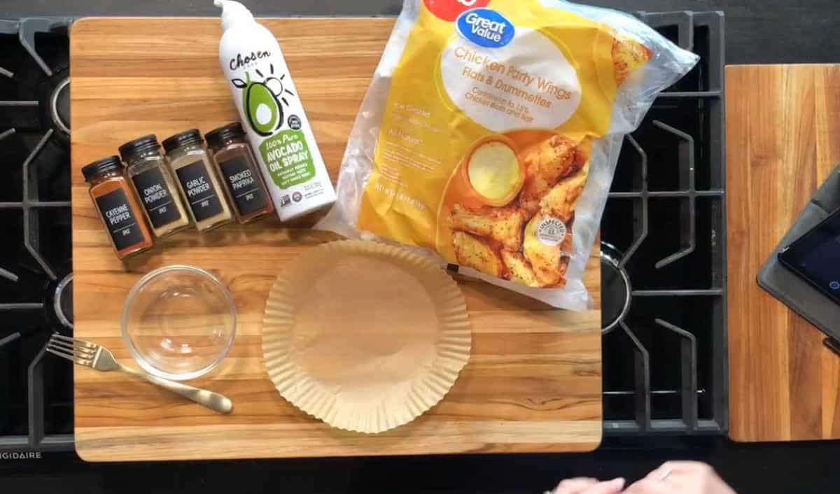 A wooden cutting board holds a pie tin with parchment paper, a small glass bowl, a fork, four spice jars, a can of avocado oil spray, and a bag of frozen chicken party wings on a stovetop.