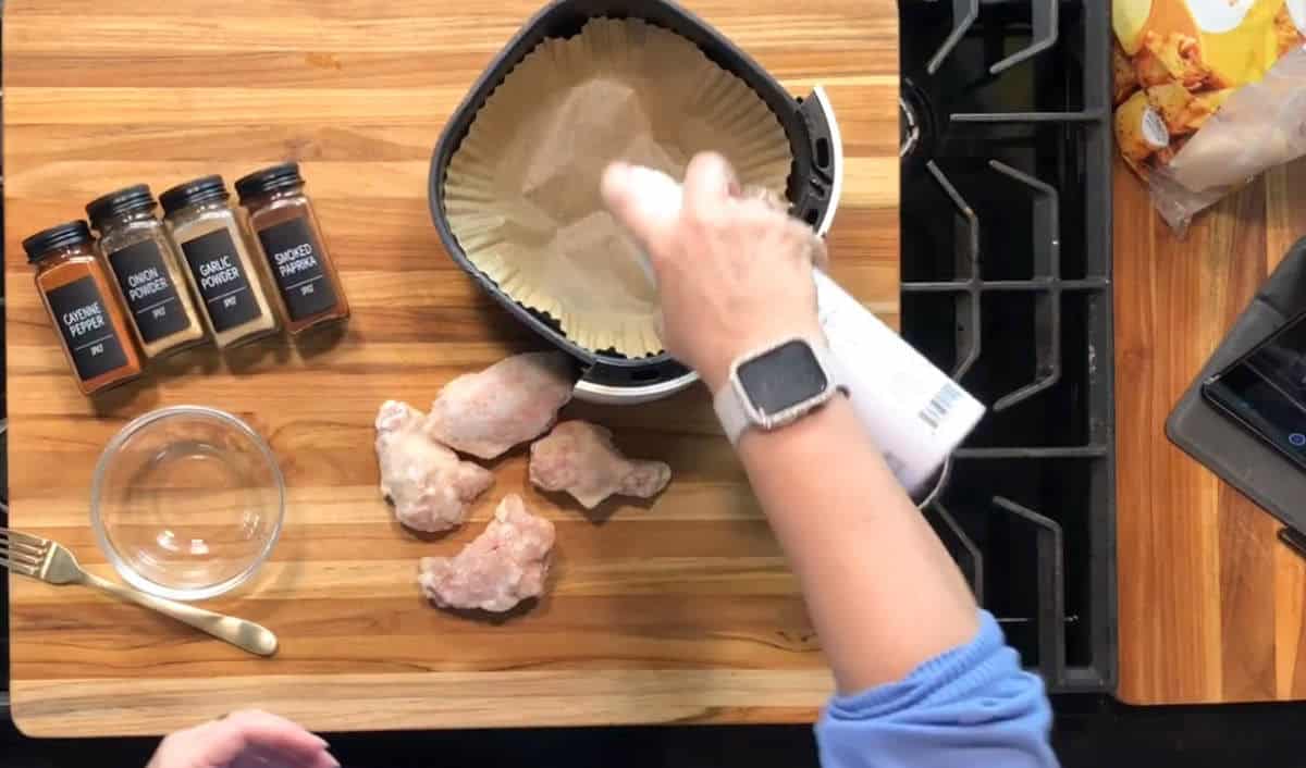 A person sprays oil over a parchment-lined air fryer basket next to four raw chicken wings, spice jars, and a bowl on a wooden countertop near a stove.