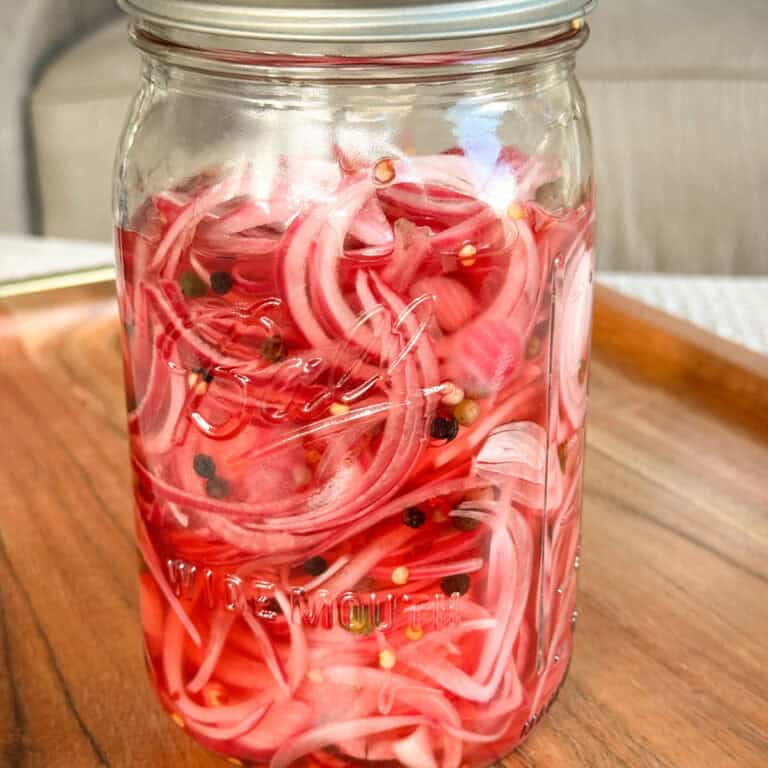 A glass mason jar filled with pickled red onions, visible thin onion slices, peppercorns, and brine, sits on a wooden tray with a metal lid on top.