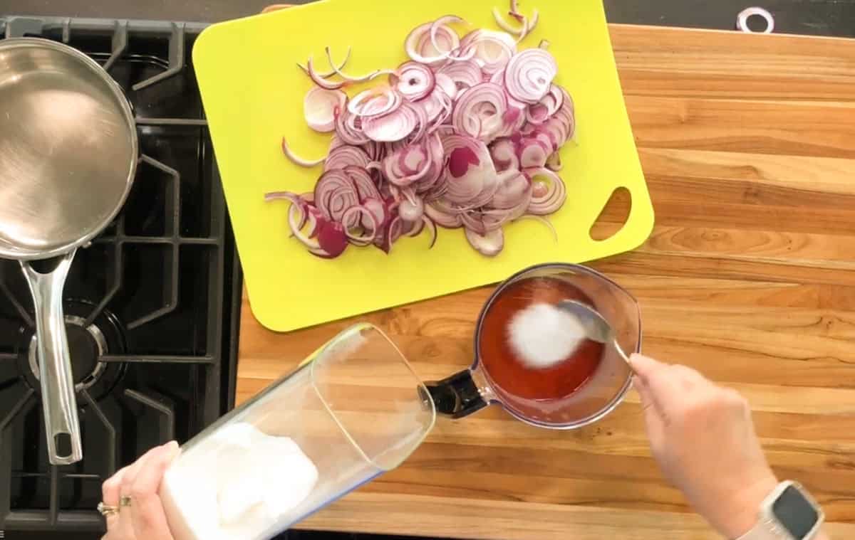 A person scoops white sugar from a container into a measuring cup of liquid, next to a cutting board with sliced red onions on a wooden counter, beside a stove and a saucepan.