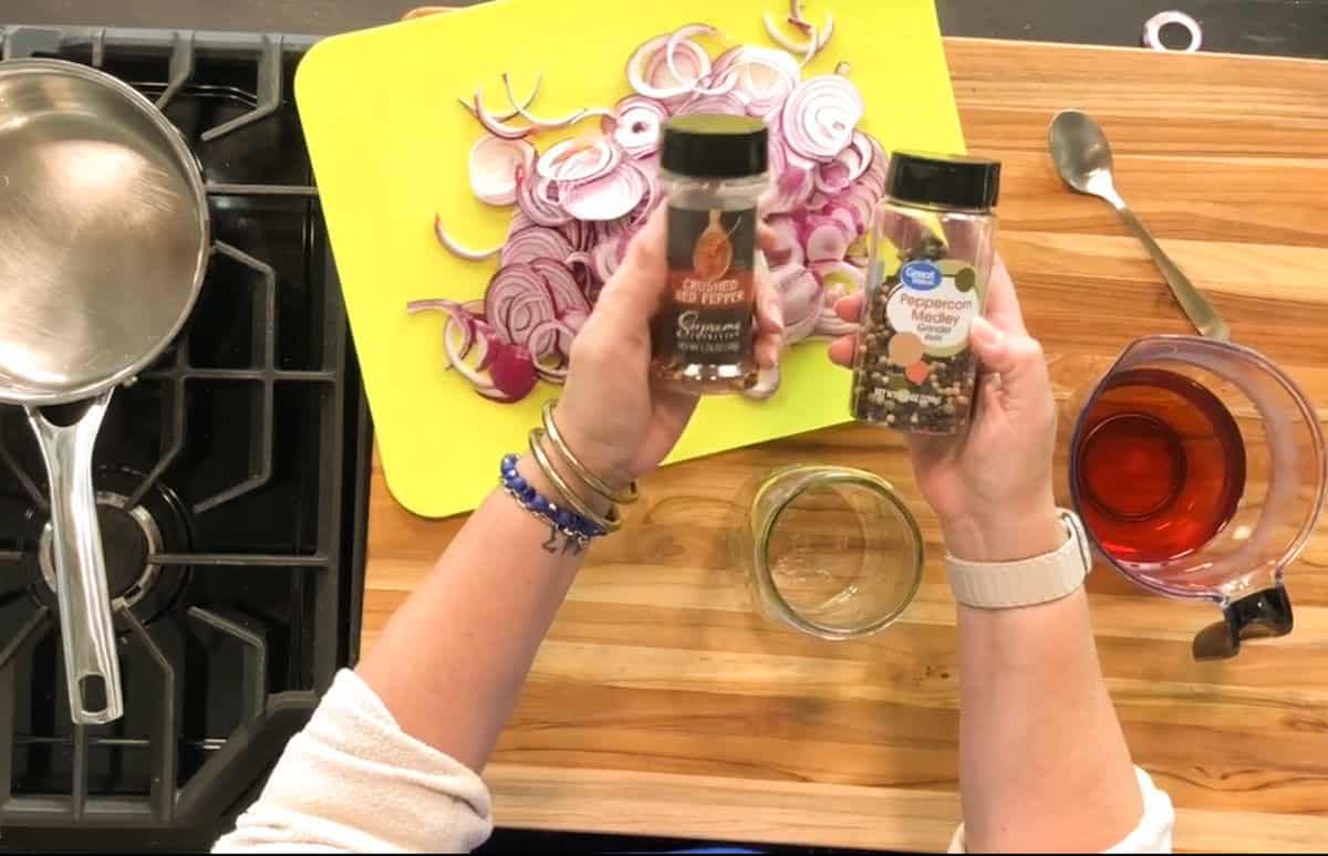 A person holds jars of spices above a cutting board with sliced red onions. A saucepan is on the stove, and a measuring cup with red liquid sits nearby on a wooden countertop.