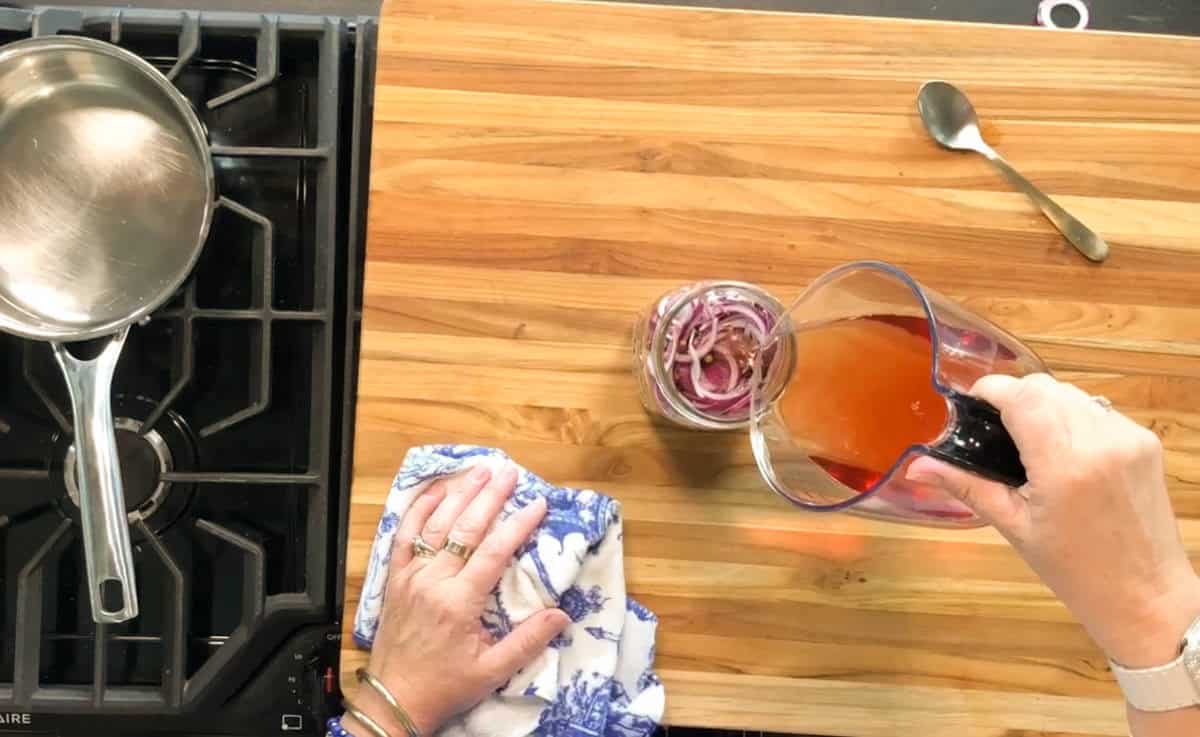 A person pours reddish liquid from a measuring cup into a jar of sliced onions on a wooden countertop. A pan is on a stove at the left, and a spoon is on the counter to the right.