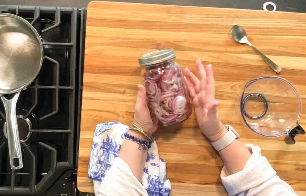 Person holding a jar of sliced red onions on a wooden cutting board, with a saucepan on a stovetop, a spoon, and a lid nearby.