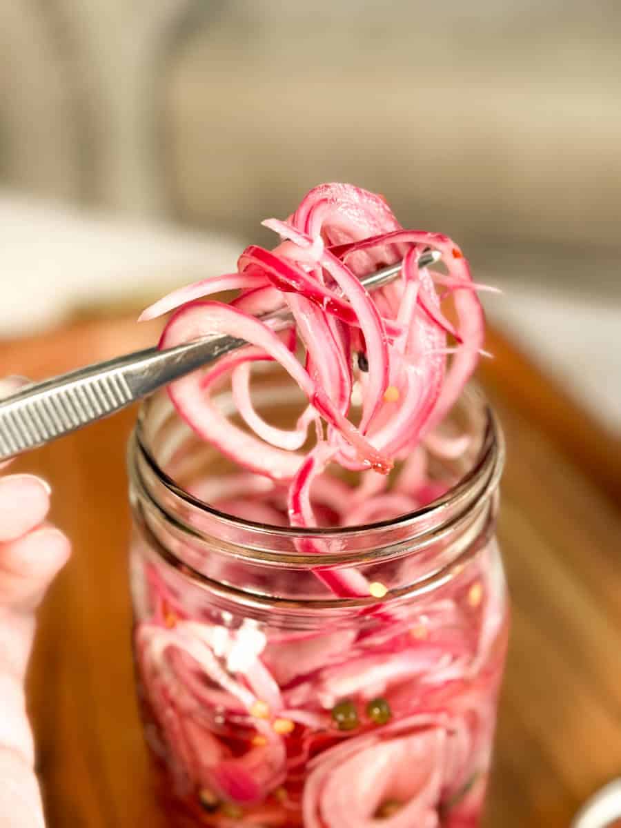 A hand holds a fork lifting pickled red onions from a glass jar filled with sliced onions and spices, placed on a wooden surface.
