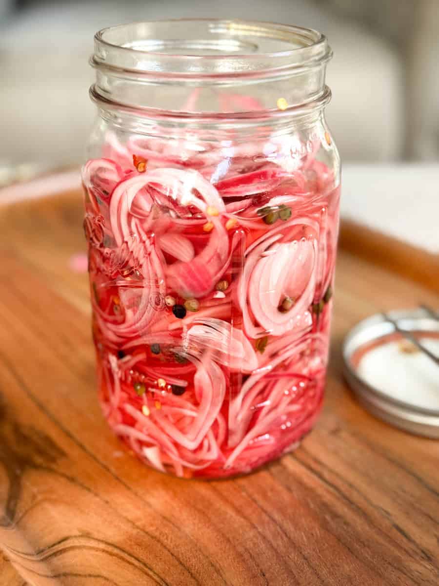 A glass jar filled with thinly sliced red onions and pickling spices, sitting on a wooden tray next to a metal lid.