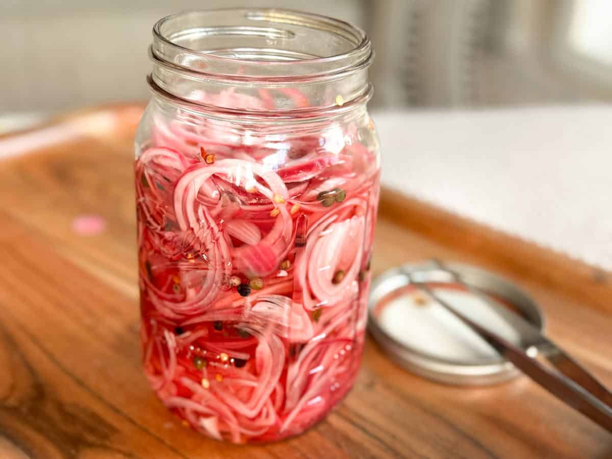 A glass jar filled with thinly sliced red onions pickling in brine, with visible spices. The jar sits on a wooden surface next to a metal lid and tongs.
