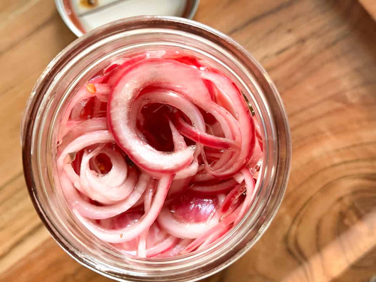 A glass jar filled with sliced red onions soaking in a pickling liquid, viewed from above on a wooden surface.