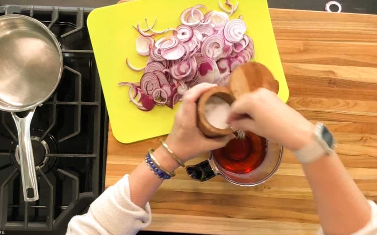 A person adds salt from a wooden container into a measuring cup filled with a red liquid next to a cutting board with sliced red onions on a wooden countertop near a stove.