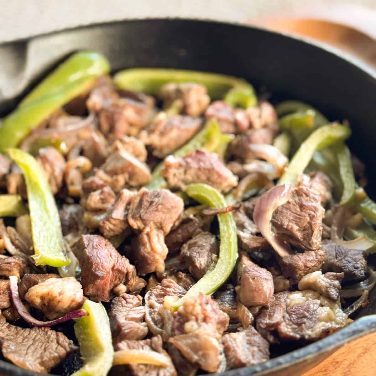 A close-up of a cast iron skillet filled with cooked beef pieces, sliced green bell peppers, and onions.
