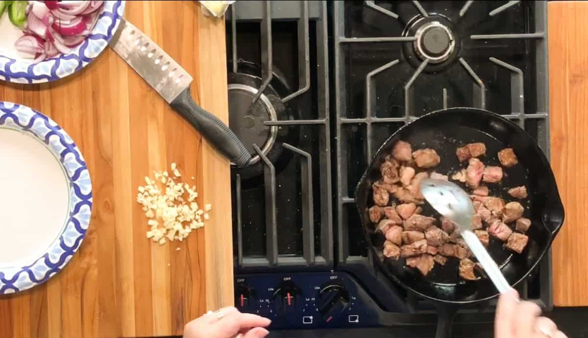 A person cooks cubed meat in a black skillet on a gas stove, stirring with a metal spoon. Chopped garlic, a knife, sliced red onions, and a paper plate are on a wooden countertop beside the stove.