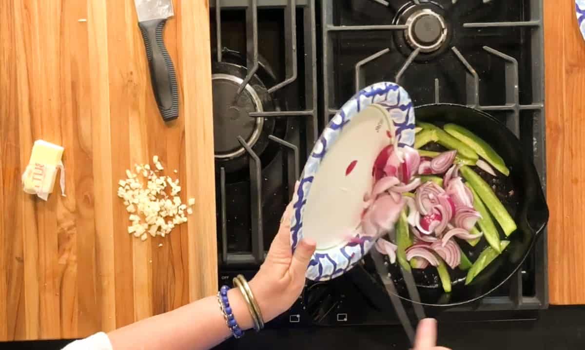 A person is adding sliced red onions from a paper plate into a skillet with green vegetables on a stovetop. Chopped garlic and a stick of butter are on a wooden counter nearby.