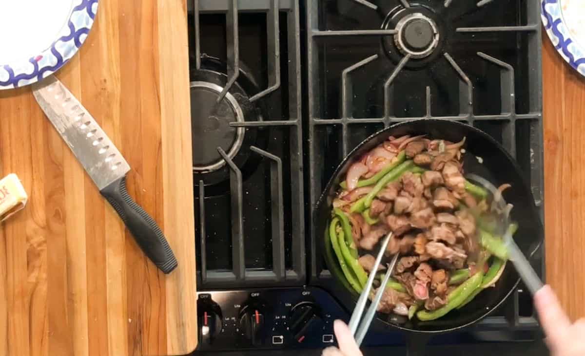 A person uses tongs to cook sliced meat, onions, and green bell peppers in a skillet on a gas stove. A chef’s knife, a stick of butter, and two plates are on the wooden countertop nearby.