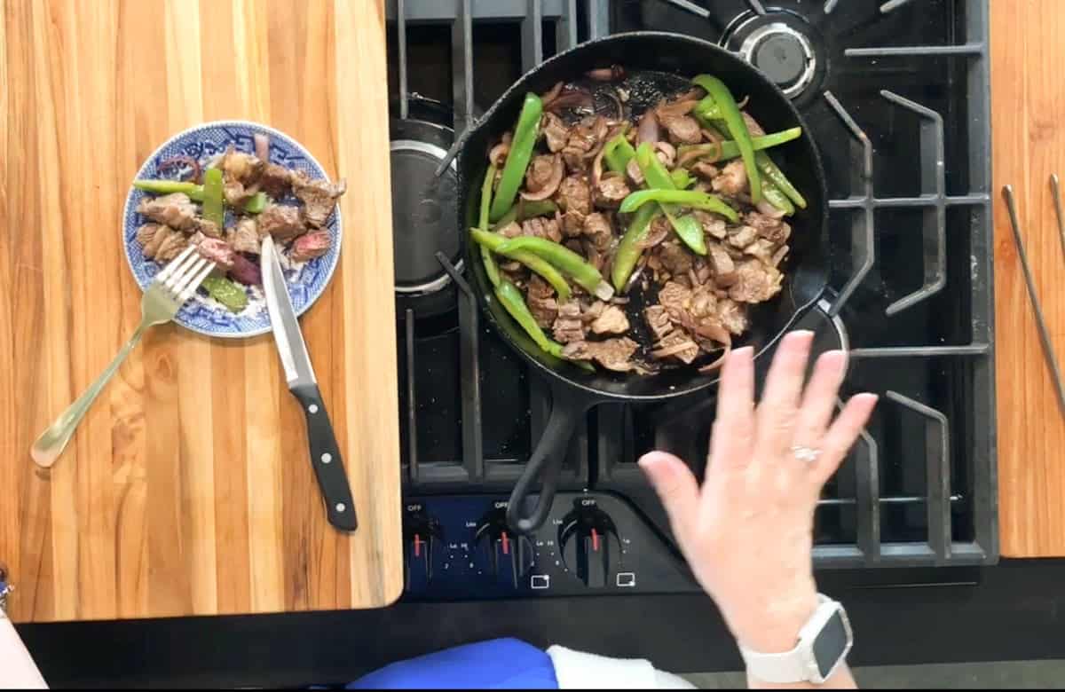 A person’s hand hovers over a cast iron skillet on a stovetop, cooking beef, green bell peppers, and mushrooms. A plate with some cooked meat, a fork, and knife sits on a wooden cutting board nearby.