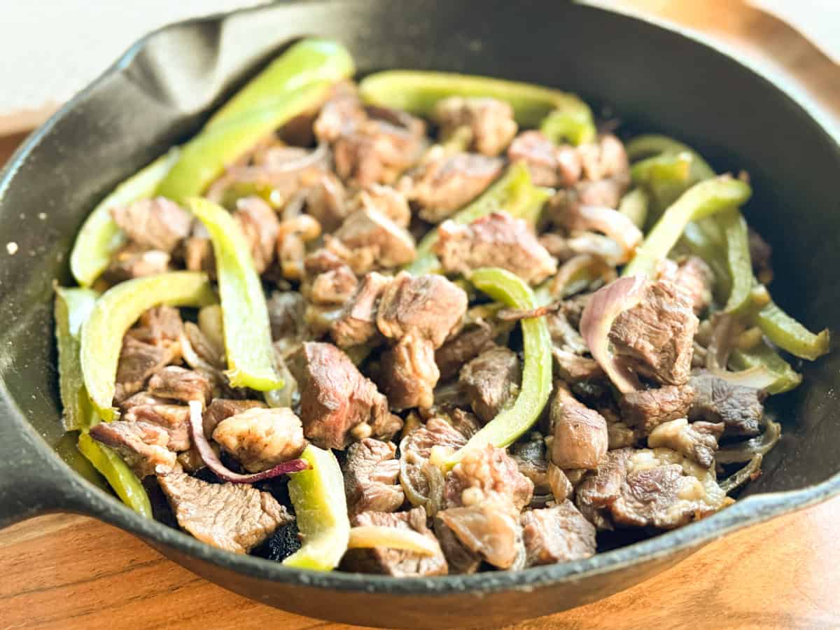 A cast iron skillet filled with cooked beef chunks, sliced green bell peppers, and onions, all sautéed together. The skillet is placed on a wooden surface.