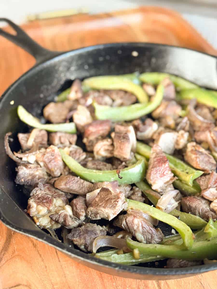 A black cast iron skillet contains cooked pieces of beef and sliced green bell peppers, placed on a wooden surface.