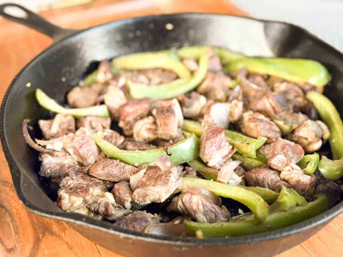 A cast iron skillet containing cooked chunks of beef and sliced green bell peppers, placed on a wooden surface.