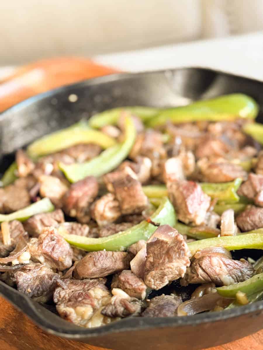 A close-up of cooked beef pieces and sliced green bell peppers in a black cast iron skillet, with a blurred background.