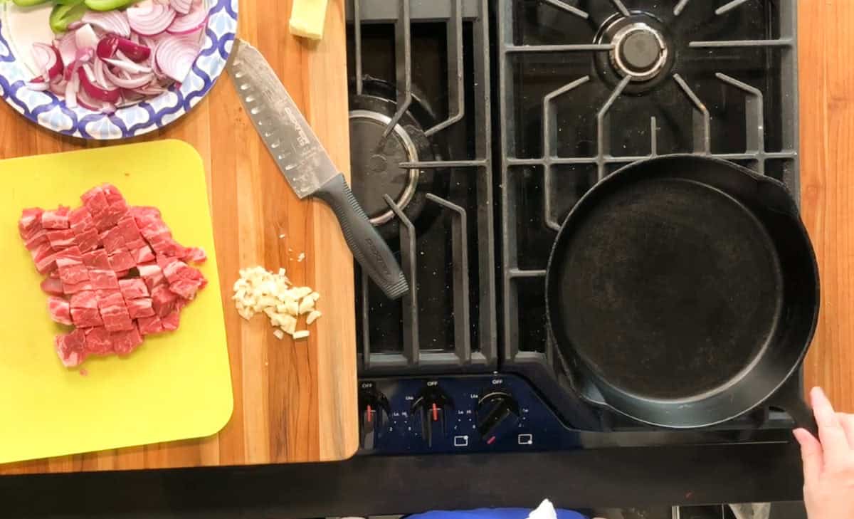 A cutting board with diced beef and chopped garlic sits next to a chef’s knife and a plate of sliced red onion and green vegetables on a wooden counter beside a stove with a black cast iron skillet.