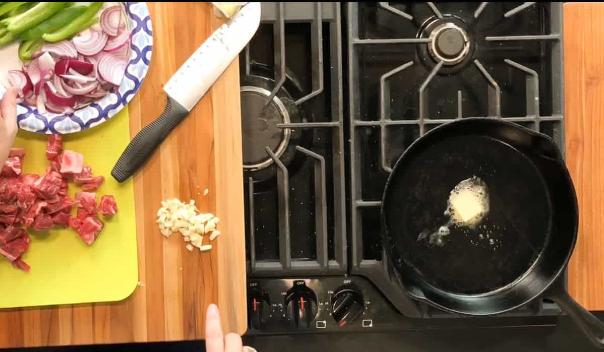 Overhead view of a kitchen counter and stove; a cast iron skillet with butter melting on a burner, chopped onions and green peppers on a plate, cubed meat on a cutting board, a pile of chopped garlic, and a knife nearby.