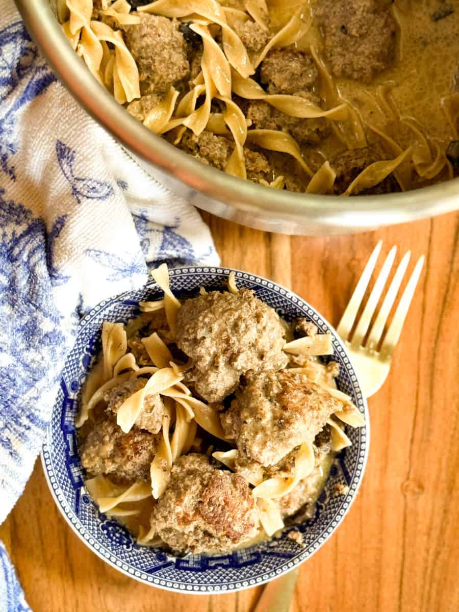 A blue and white bowl filled with egg noodles and meatballs sits on a wooden surface next to a gold fork, with a larger pot of the same dish and a blue patterned towel beside it.