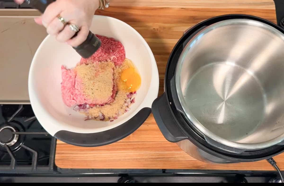 A hand is grinding pepper into a bowl containing ground meat, an egg, breadcrumbs, and chopped onions. An empty stainless steel pressure cooker sits nearby on a wooden countertop next to a stovetop.
