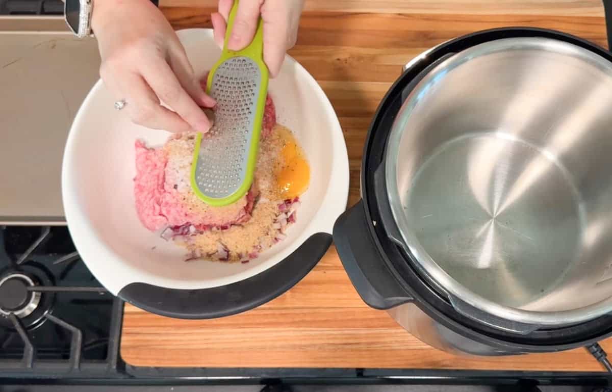 A person grates an ingredient into a bowl containing ground meat, breadcrumbs, chopped onions, and an egg. An empty pressure cooker sits on a wooden countertop next to the bowl.