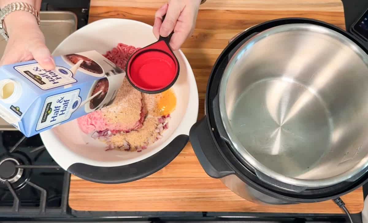 A person pours a carton of half & half into a measuring cup over a bowl containing ground meat, an egg, breadcrumbs, and seasonings next to an open Instant Pot on a wooden countertop.