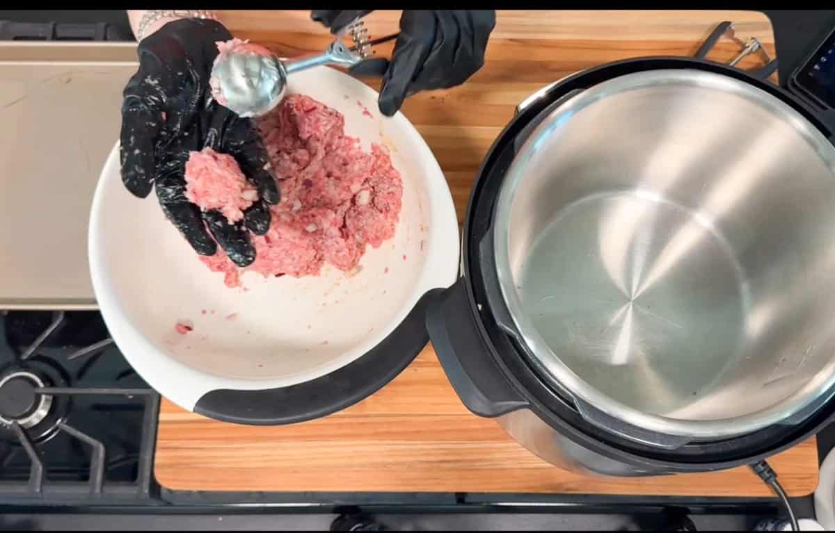 A person wearing black gloves scoops raw ground meat from a white bowl with a metal scoop, next to an empty stainless steel pot on a wooden countertop.