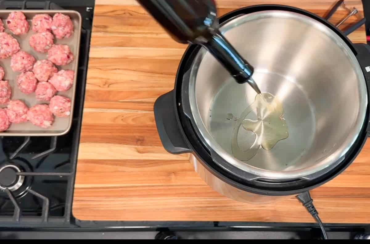 A person pours oil from a bottle into an electric pressure cooker on a wooden countertop; a metal tray of raw meatballs sits nearby next to a stovetop.