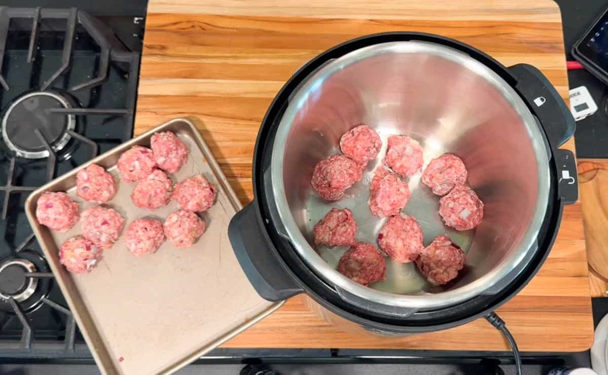 Raw meatballs are arranged inside an Instant Pot, with more uncooked meatballs on a baking sheet nearby. The scene is set on a wooden countertop next to a gas stove.