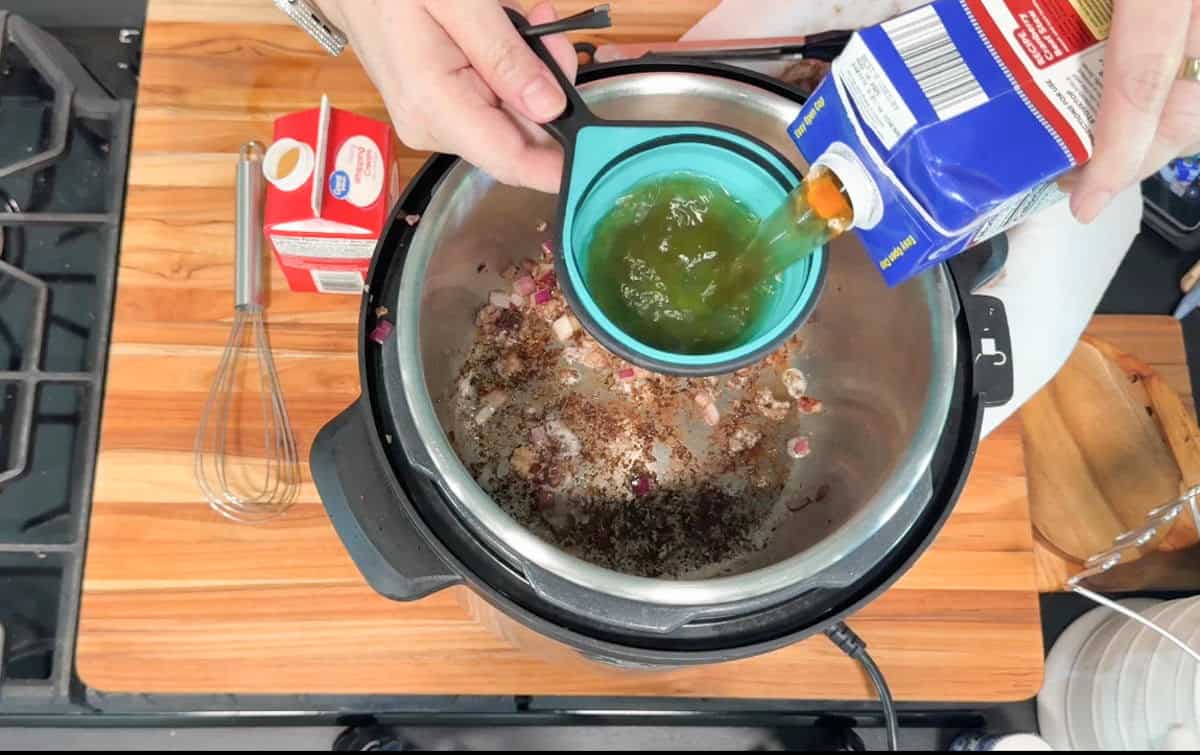 A person pours broth from a carton into a blue measuring cup over an electric pressure cooker that contains chopped onions and seasonings. A whisk, a smaller carton, and a cutting board are on the wooden countertop nearby.