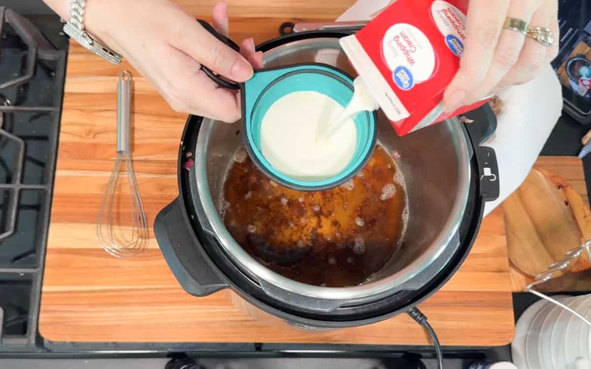 A person pours heavy whipping cream from a carton into a measuring cup over an open Instant Pot filled with a brown liquid. A whisk and various utensils are on a wooden countertop beside the pot.