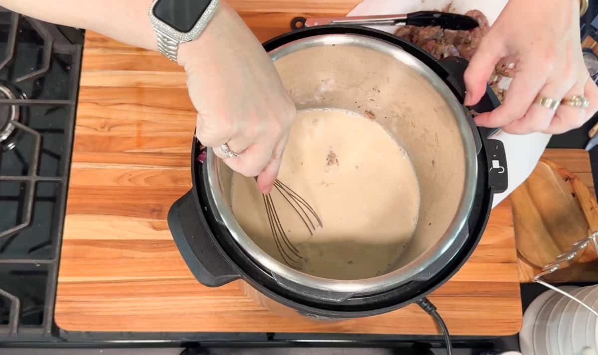 A person uses a whisk to stir a creamy mixture inside an Instant Pot on a wooden countertop, with a stove visible to the left and food on a cutting board to the right.