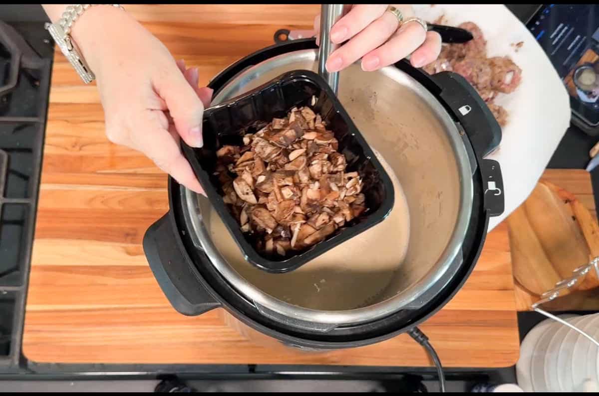 A person holding a black plastic container of chopped mushrooms over an open pressure cooker on a wooden countertop, preparing to add them to the pot.
