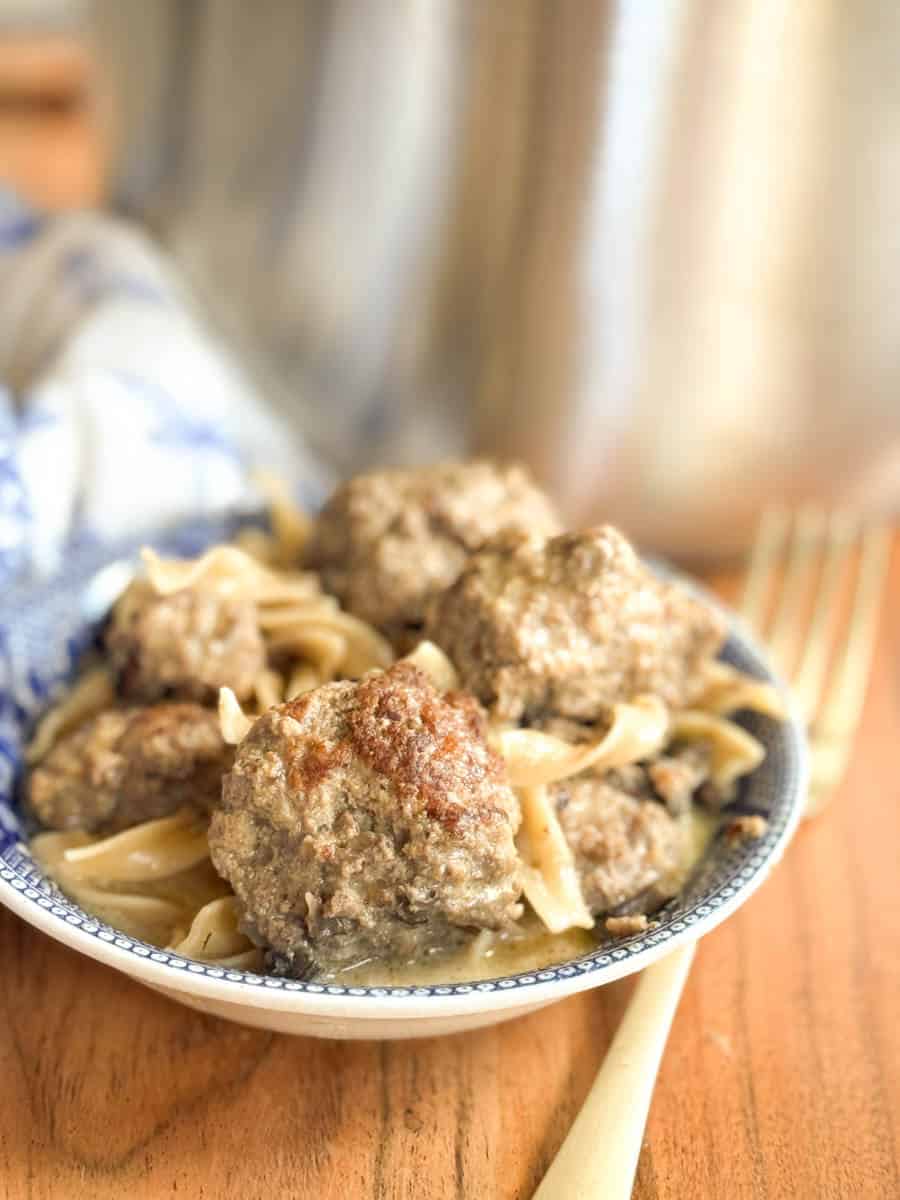 A close-up of a plate with several meatballs served over egg noodles, with a fork placed next to the plate on a wooden surface. A blue and white cloth is partially visible in the background.