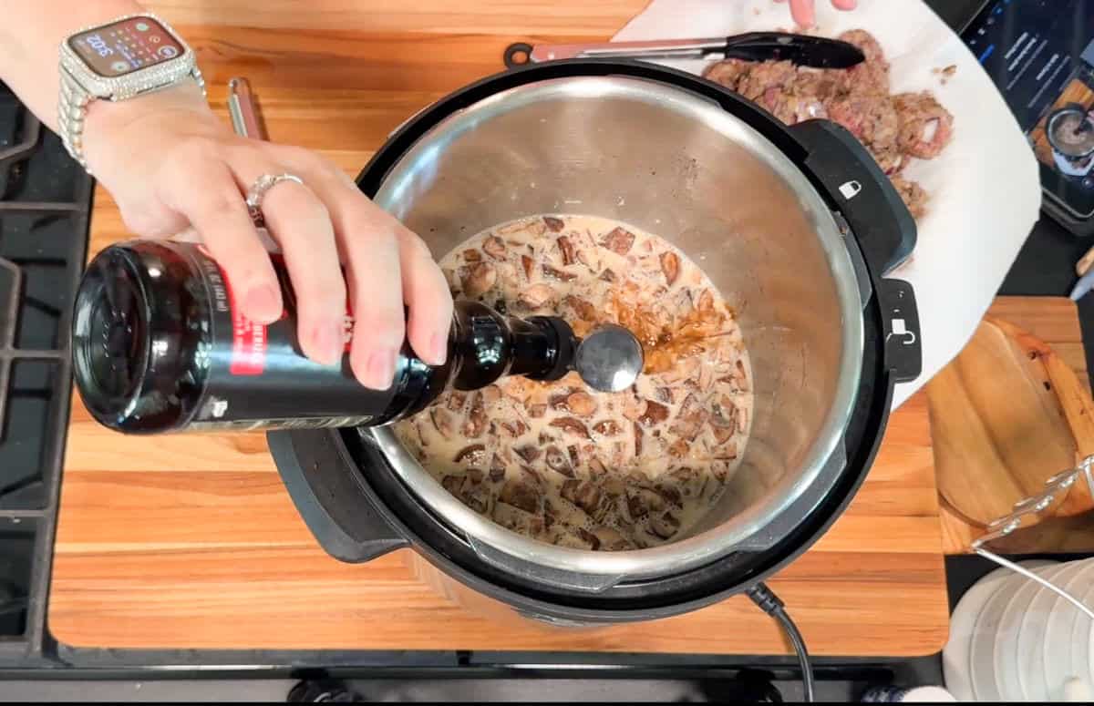 A person pours a dark liquid from a bottle into an electric pressure cooker filled with a creamy mixture and mushrooms. Cooked meat rests on a plate lined with paper towels nearby. A watch is visible on the person’s wrist.