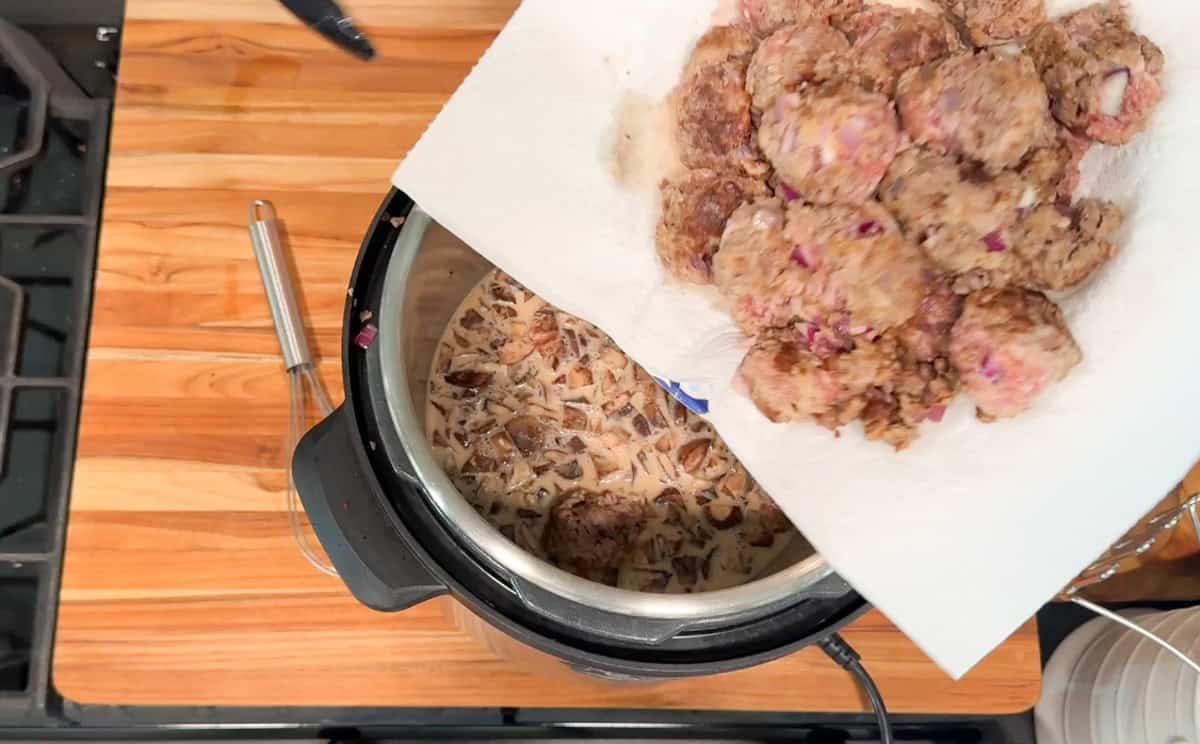 A hand holds a plate with raw meatballs above an open pressure cooker containing chopped mushrooms and liquid on a wooden countertop next to a stove.
