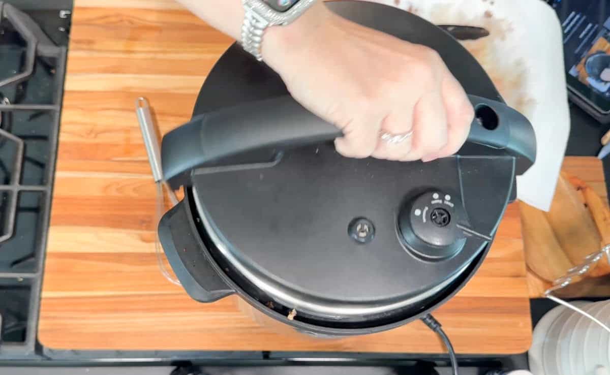 A hand is opening the lid of an electric pressure cooker on a wooden countertop. Part of a stovetop, a cutting board, and a plate with food scraps are visible nearby.