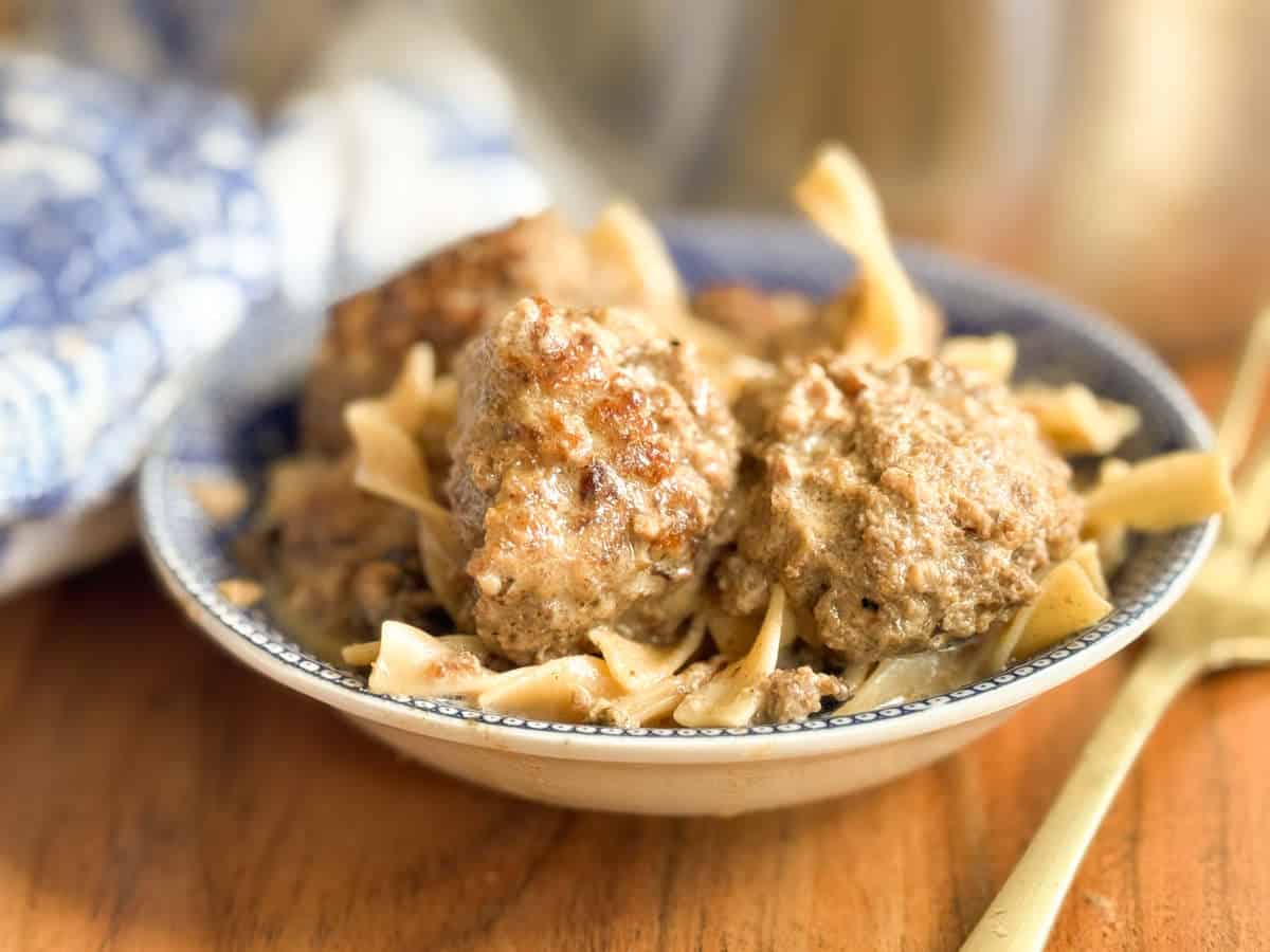 A ceramic bowl with two browned meatballs on top of wide egg noodles in a creamy sauce, placed on a wooden table beside a gold-colored fork and a blue-and-white patterned cloth.